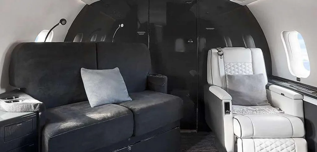 A close-up view of the private jet cabin, featuring a dark grey sofa with pillows and a white quilted seat.