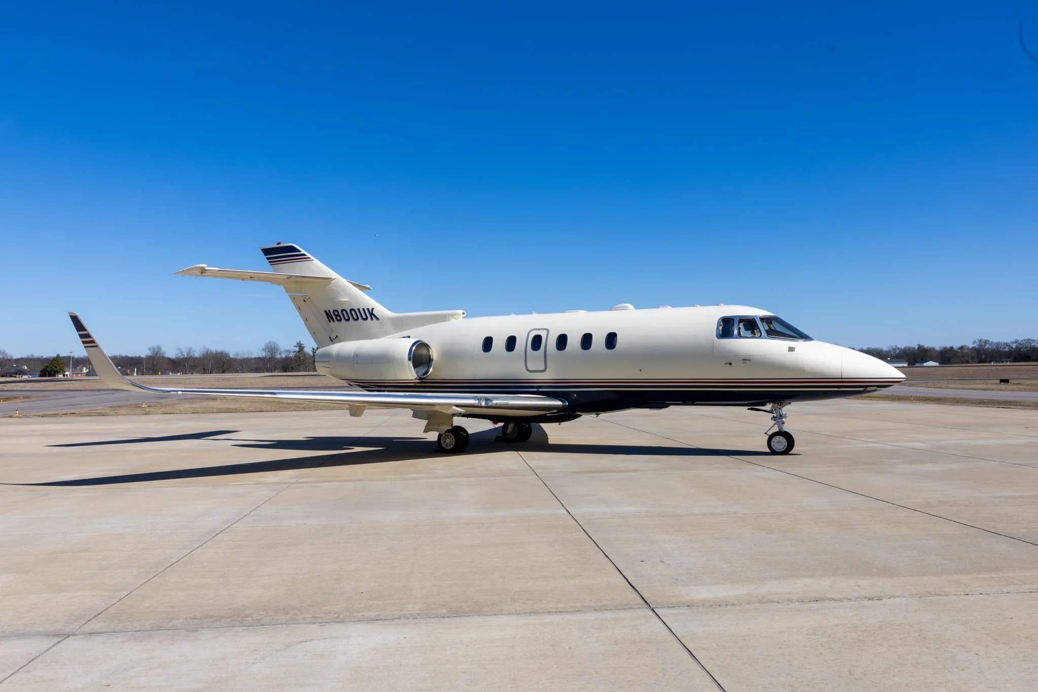 A white and blue business jet, N800UK, is parked on a concrete tarmac under a clear blue sky.