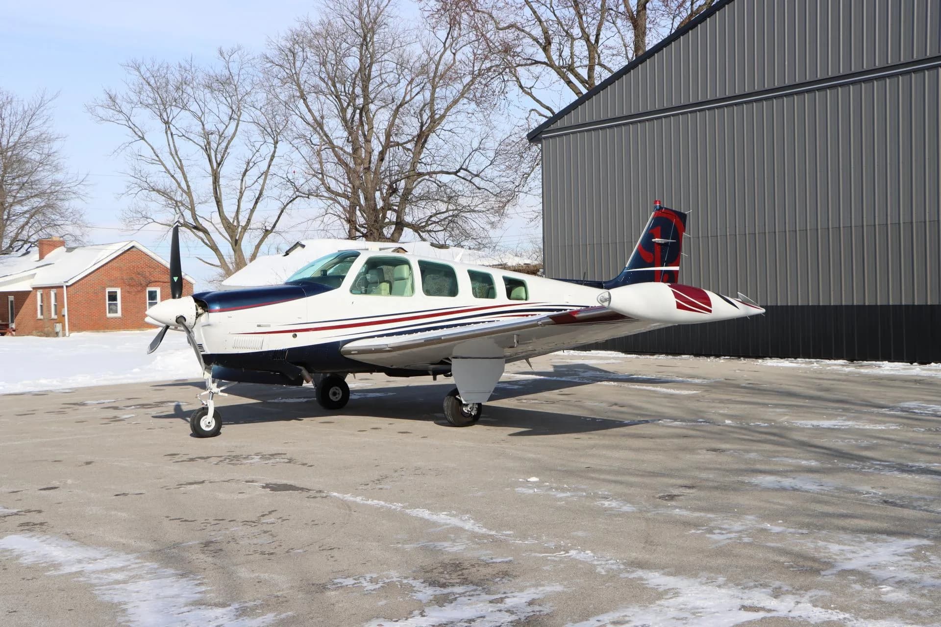 Side view of a white, blue, and red single-engine aircraft parked on a partially snowy tarmac next to a hangar.