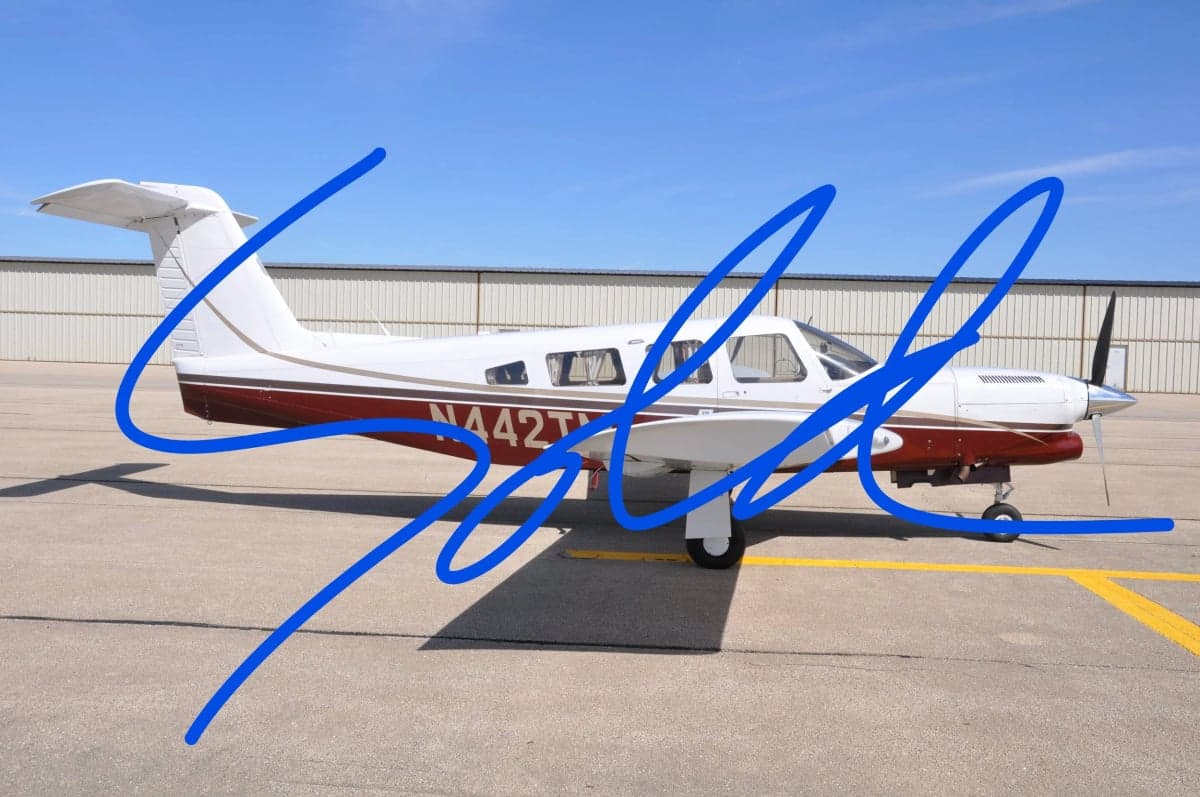 Side view of a Piper aircraft with a white and maroon paint scheme, parked on the tarmac with a blue scribble overlay.