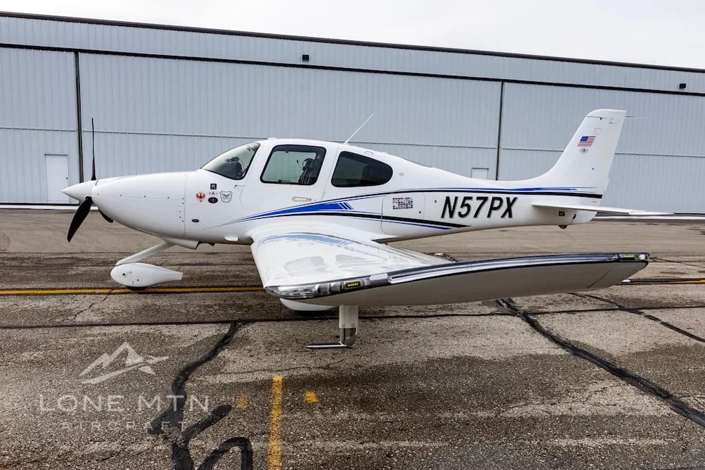 A side view of a white Cirrus SR22 aircraft with blue and black stripes, tail number N57PX, parked on a wet airport tarmac.