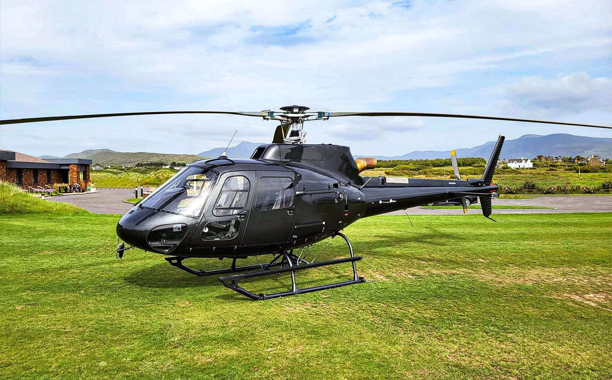 A sleek black helicopter, likely an Airbus H125, is parked on a vibrant green lawn with a modern building and distant mountains under a cloudy sky.