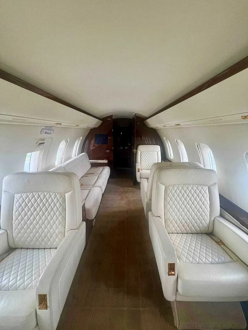 The luxurious interior cabin of a private jet, featuring quilted white leather seats and a brown carpeted aisle.