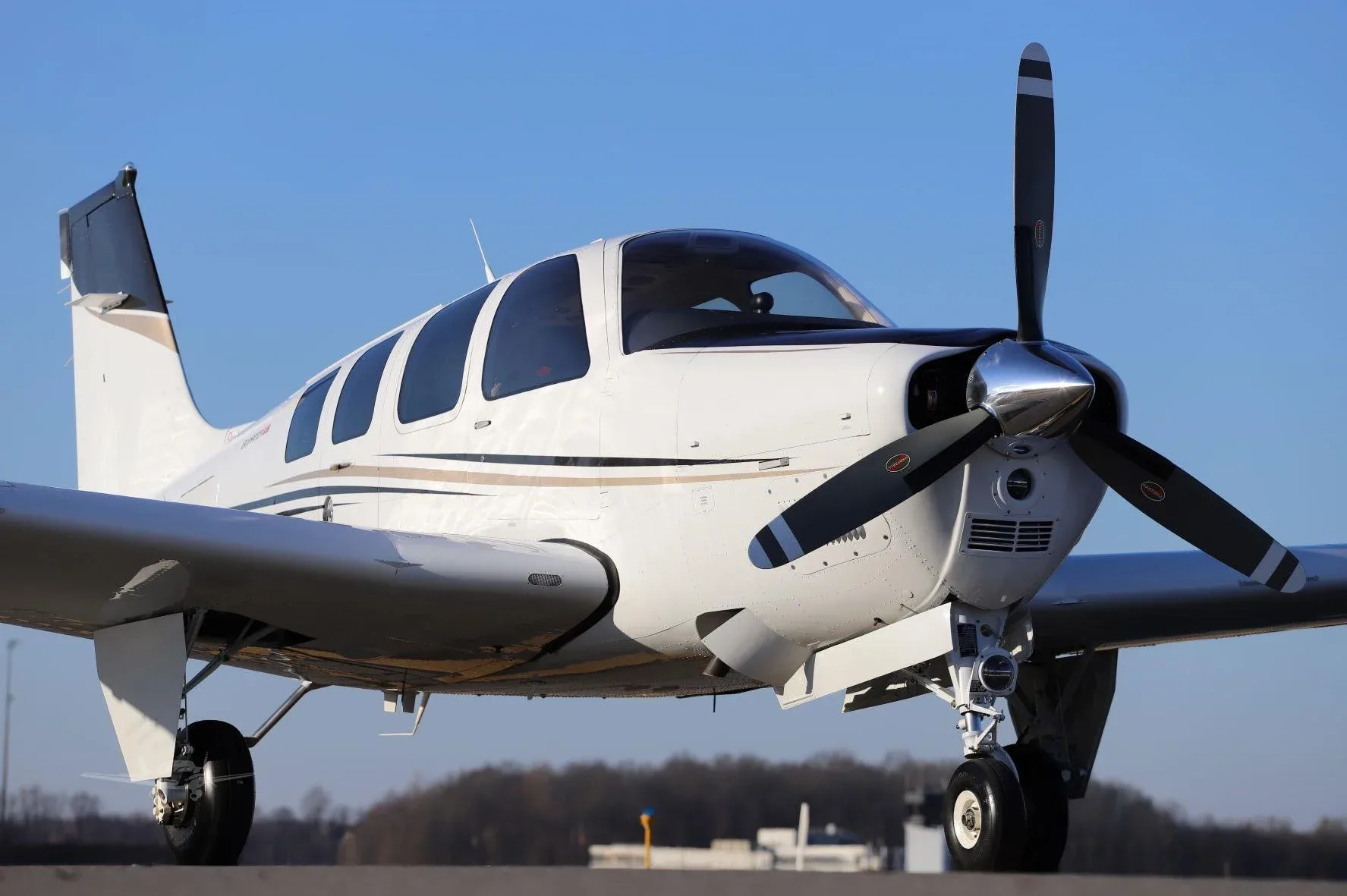 A white Beechcraft Bonanza G36 with black and gold stripes, seen from the front-right on a sunny day.