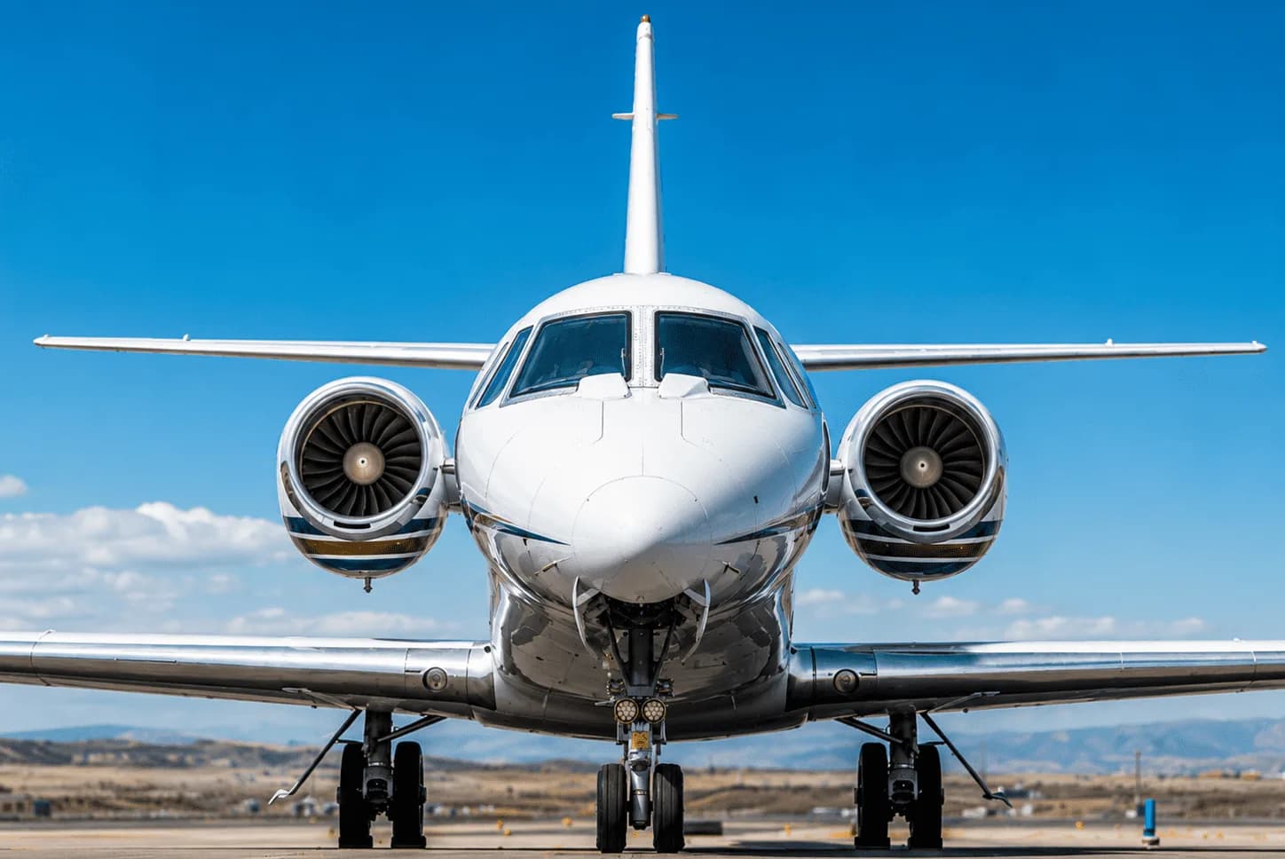 A white business jet with dark accent stripes, viewed head-on on a tarmac under a clear blue sky.