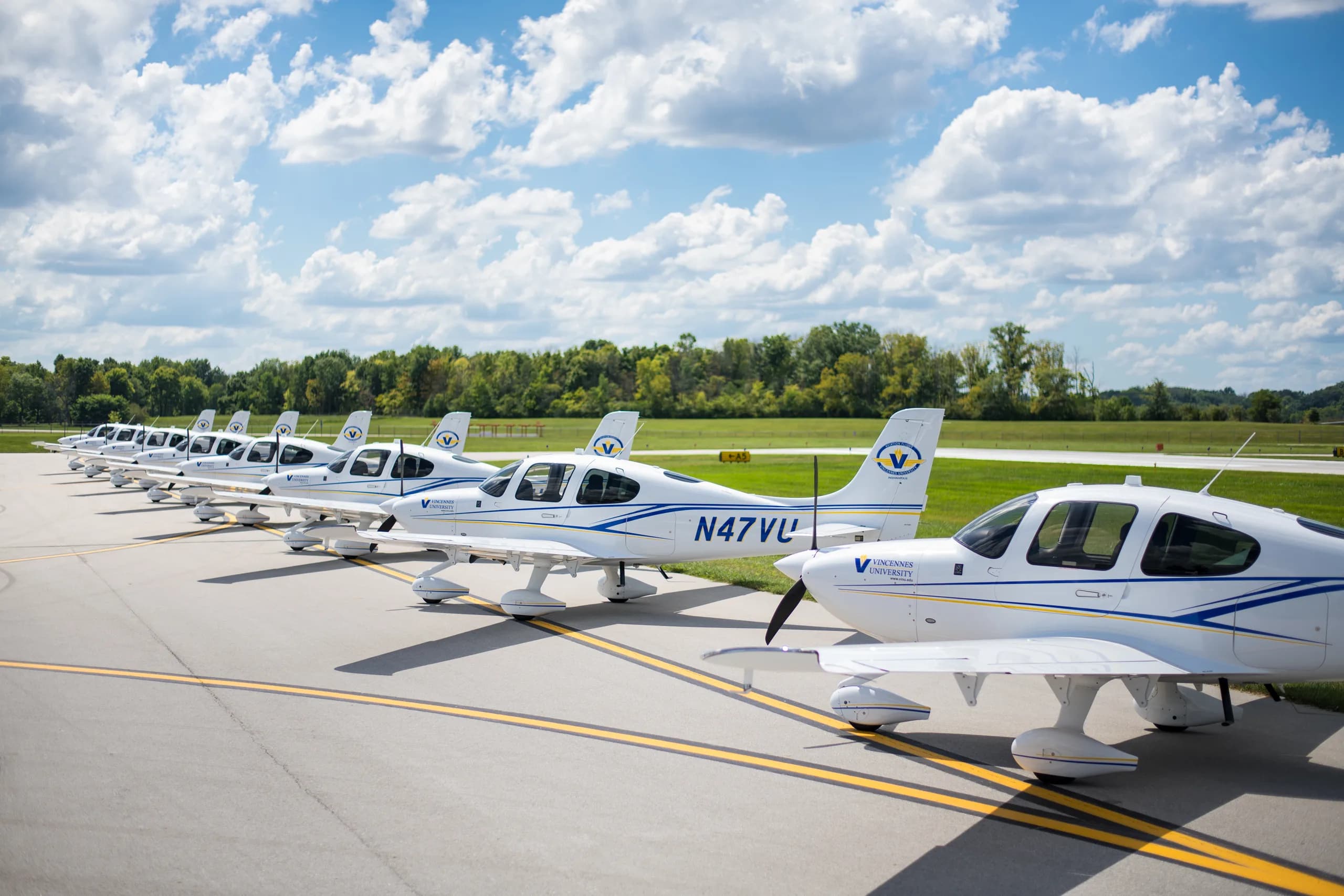 A line of white single-engine propeller aircraft with blue and yellow stripes and "Vincennes University" logos, parked on an asphalt runway under a blue sky with clouds.
