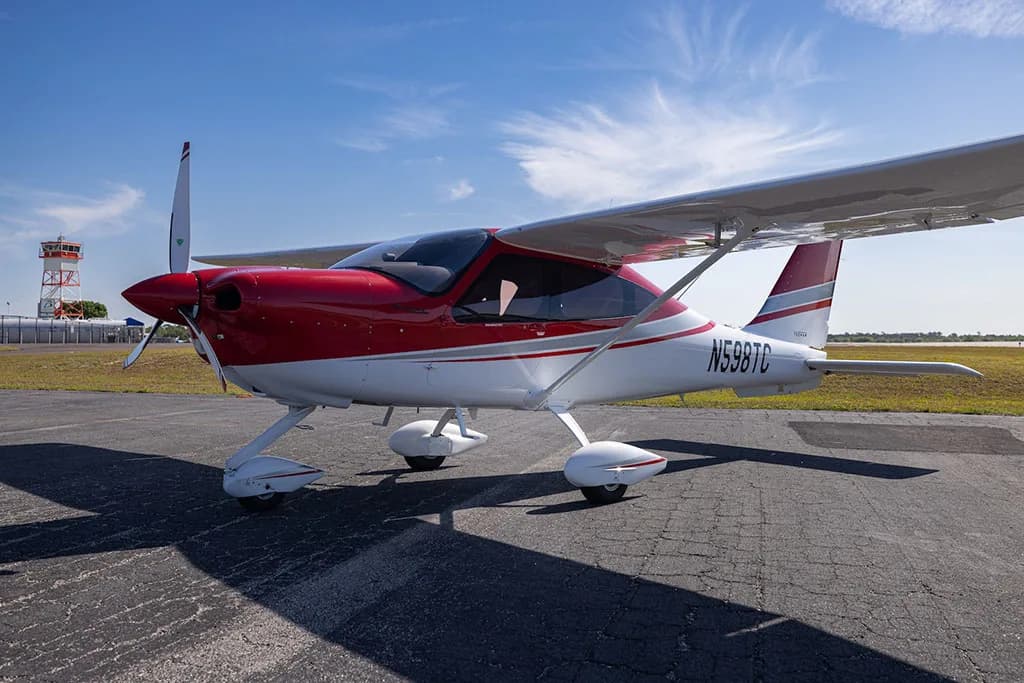 A red and white Tecnam P2010 aircraft with registration N598TC parked on an airfield under a blue sky.