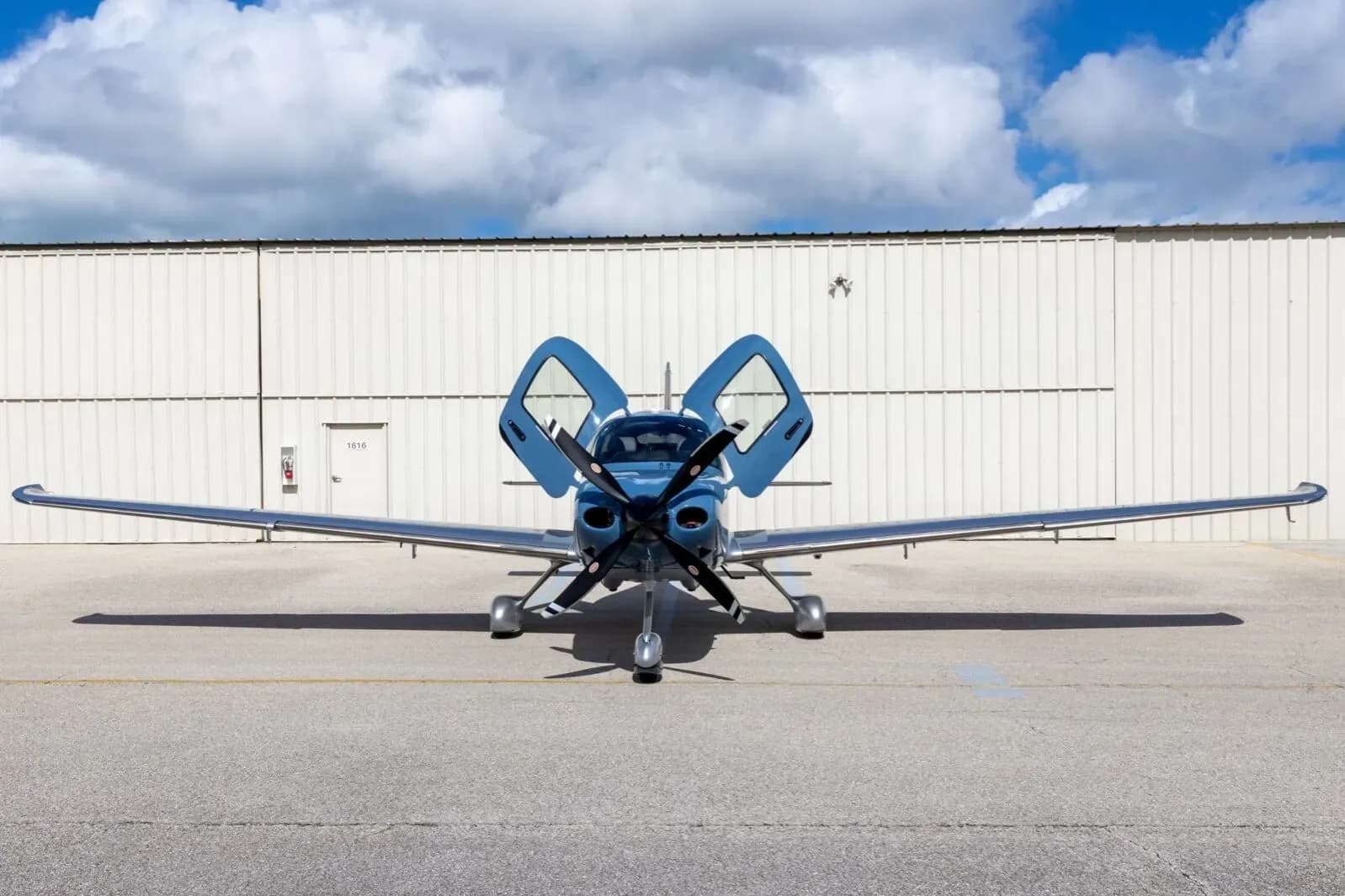 A front view of a blue and silver Cirrus SR22T aircraft with its gull-wing doors open, parked in front of a white hangar.