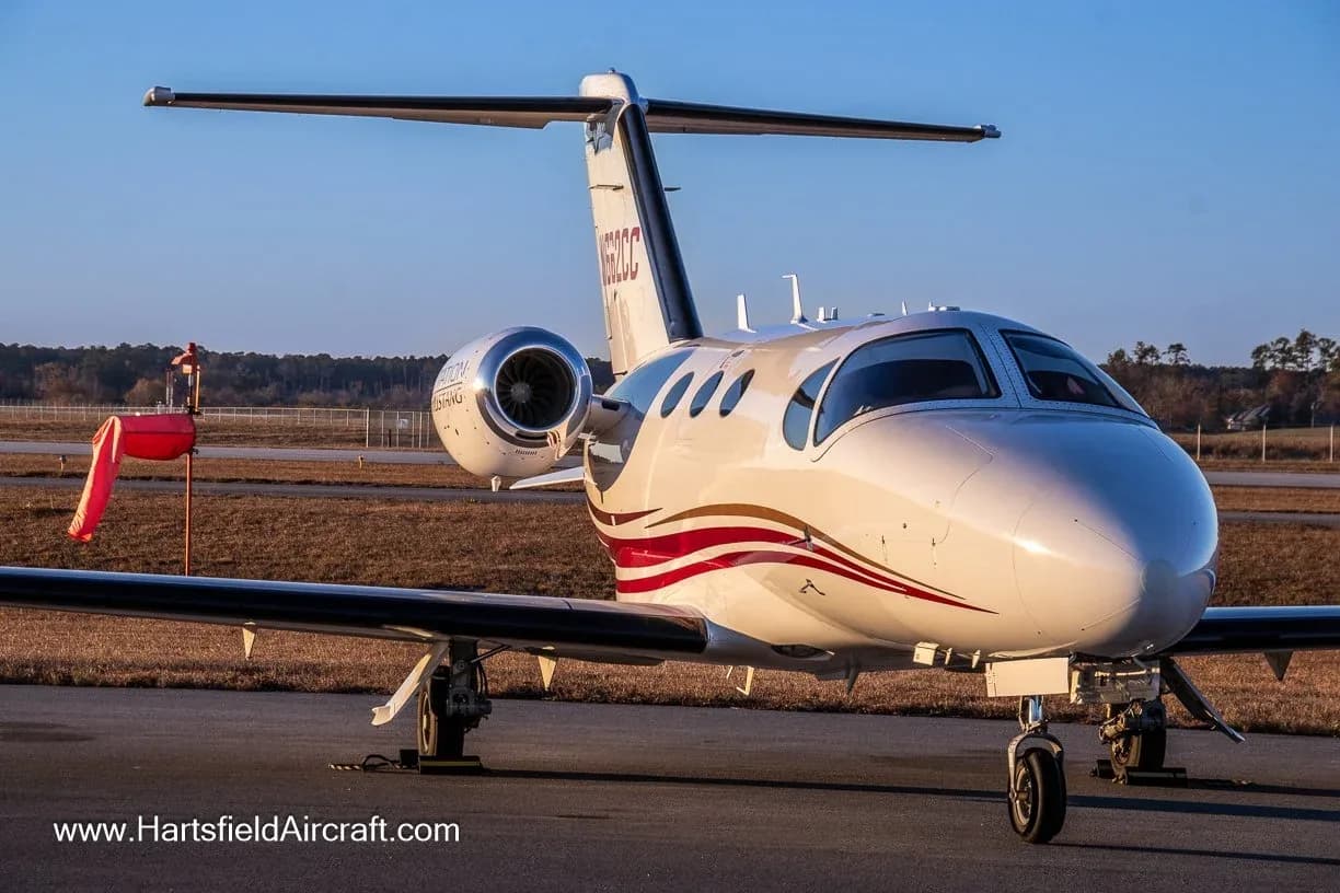 A white Cessna Citation Mustang business jet with red and gold stripes is parked on an asphalt ramp at an airfield under a clear blue sky.