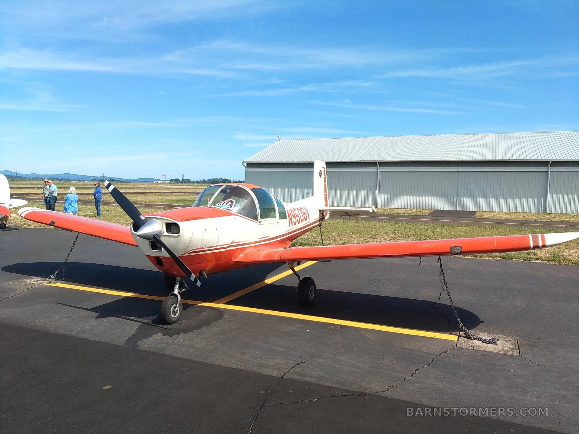 A red and white single-engine aircraft, N9506V, is parked on the tarmac in front of a hangar.