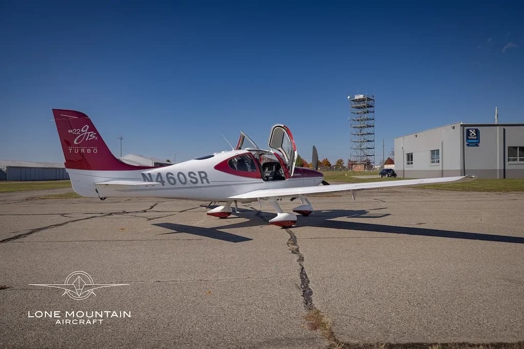 A white and maroon Cirrus SR22T aircraft, N460SR, with open doors, parked on an airfield with a control tower in the background.