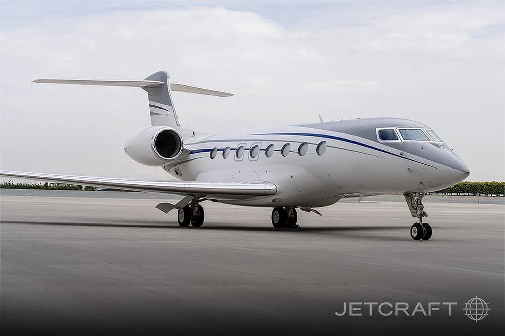 A Gulfstream G650 business jet with a white and grey paint scheme, parked on an airfield under a cloudy sky.