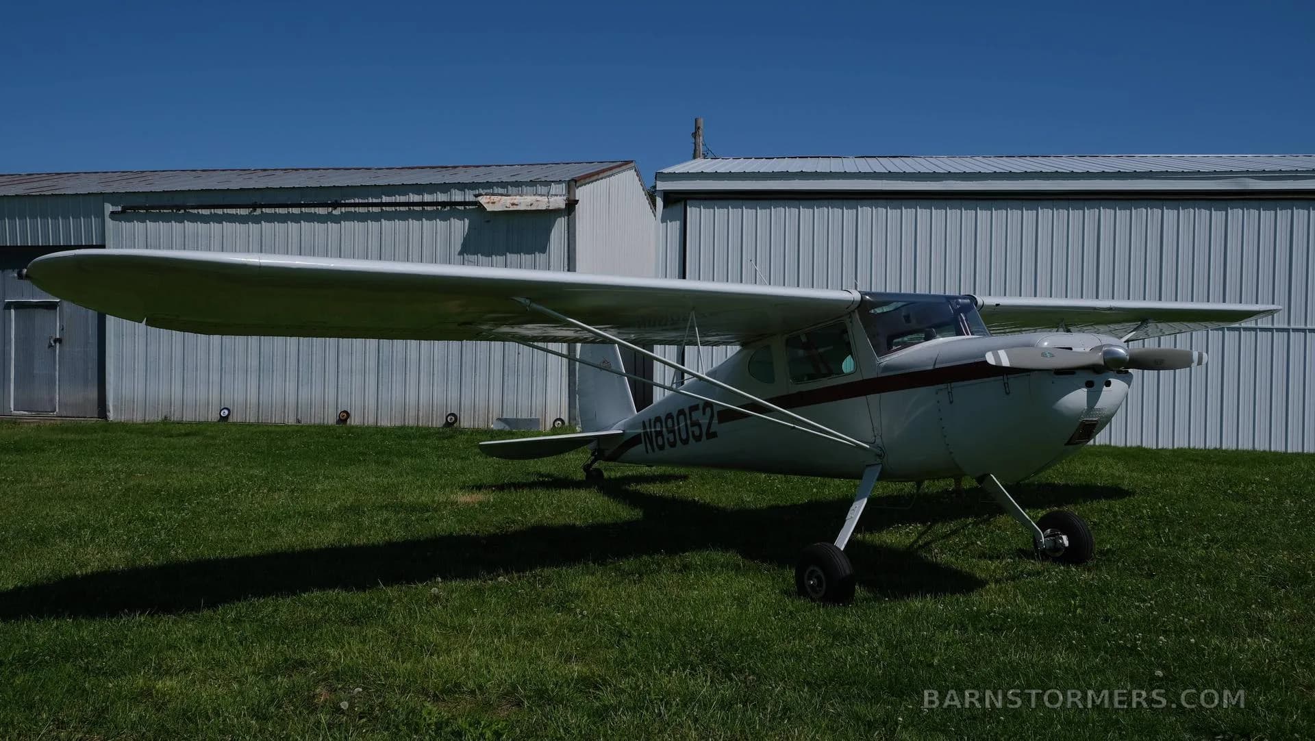 A white Cessna 170 with a red stripe, registration N89052, parked on green grass in front of hangars.