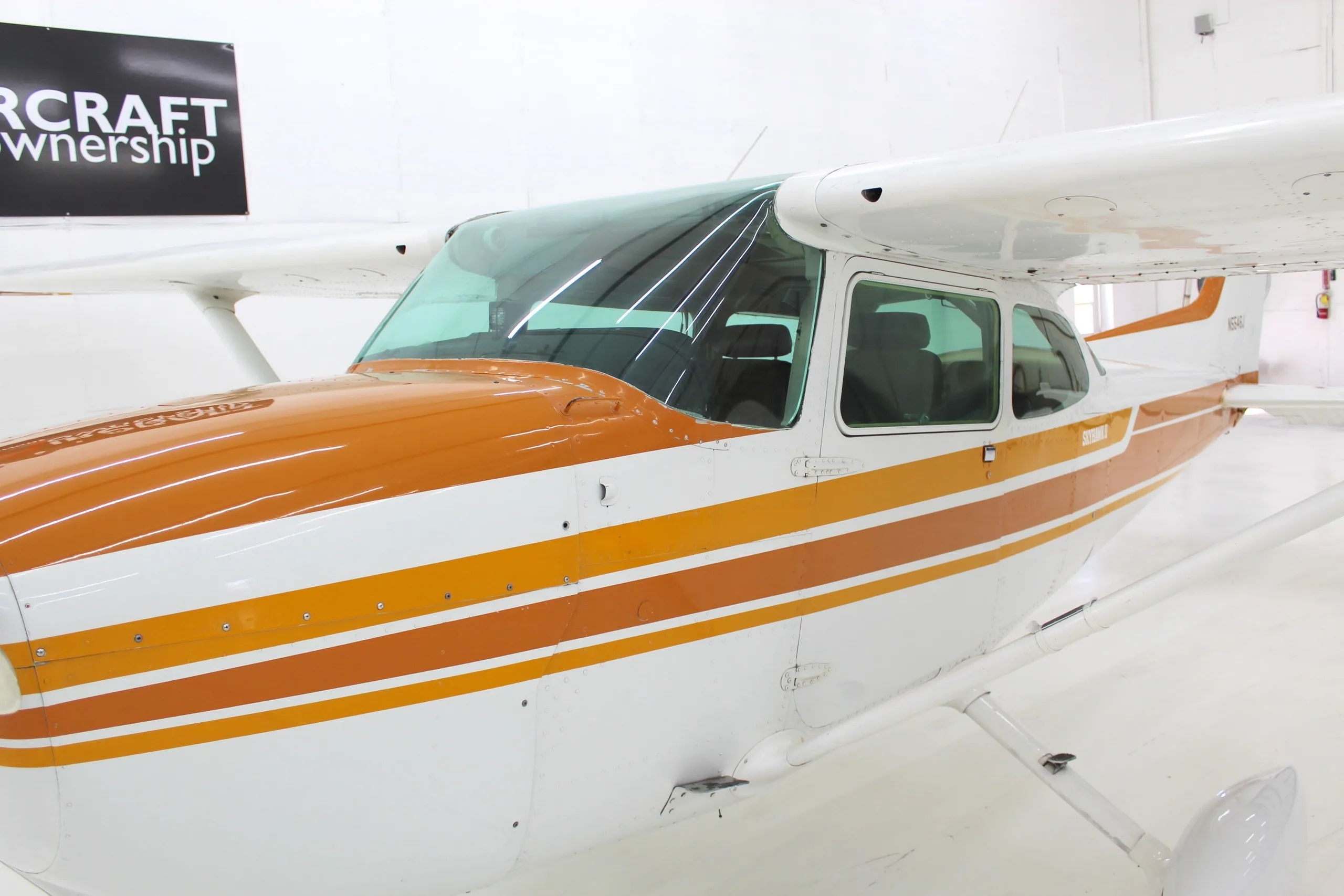 A close-up view of the front fuselage and wing of a white and orange Cessna Skyhawk II (N5546J) in a hangar.