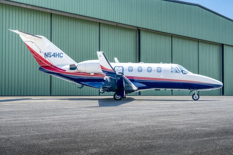 A white, blue, and red Cessna Citation jet (N54HC) is parked on an asphalt tarmac in front of a green hangar.