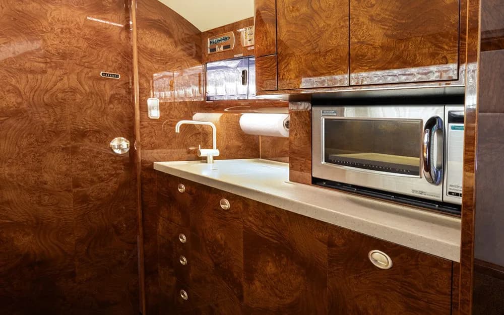 A compact galley in a private jet, equipped with a microwave, sink, and wood cabinetry.