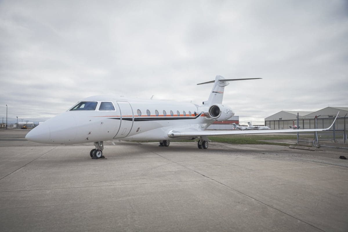 A white private jet is parked on the tarmac with a hangar in the background.