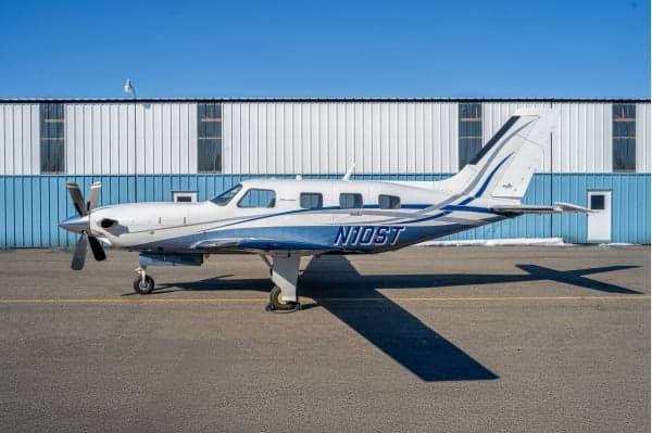 A white and blue Piper M600 single-engine turboprop aircraft is parked on an tarmac in front of a hangar.