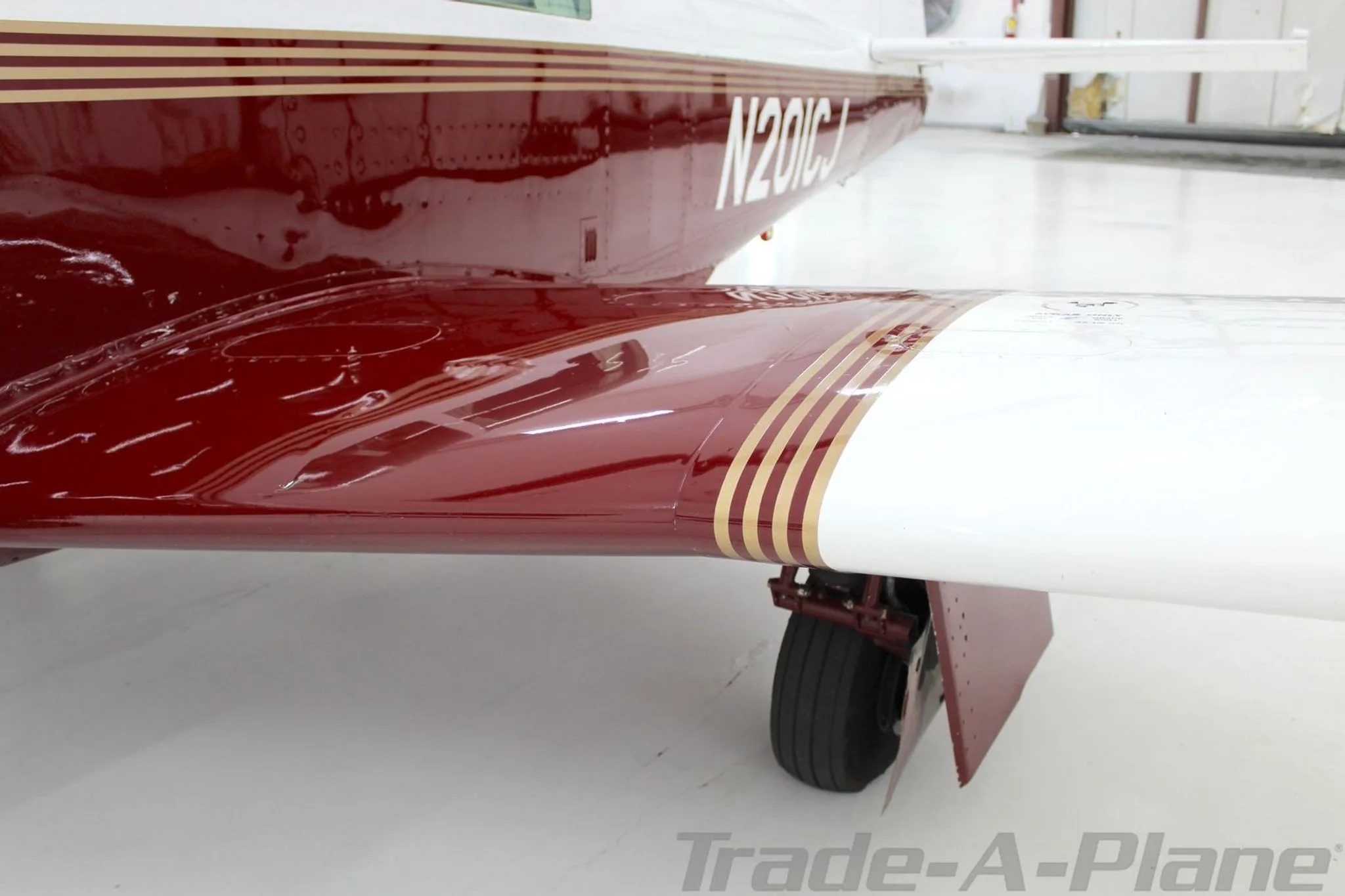 A close-up view of the wing root and fuselage of the Mooney M20J, N201CJ, highlighting the maroon and white paint with gold stripes.