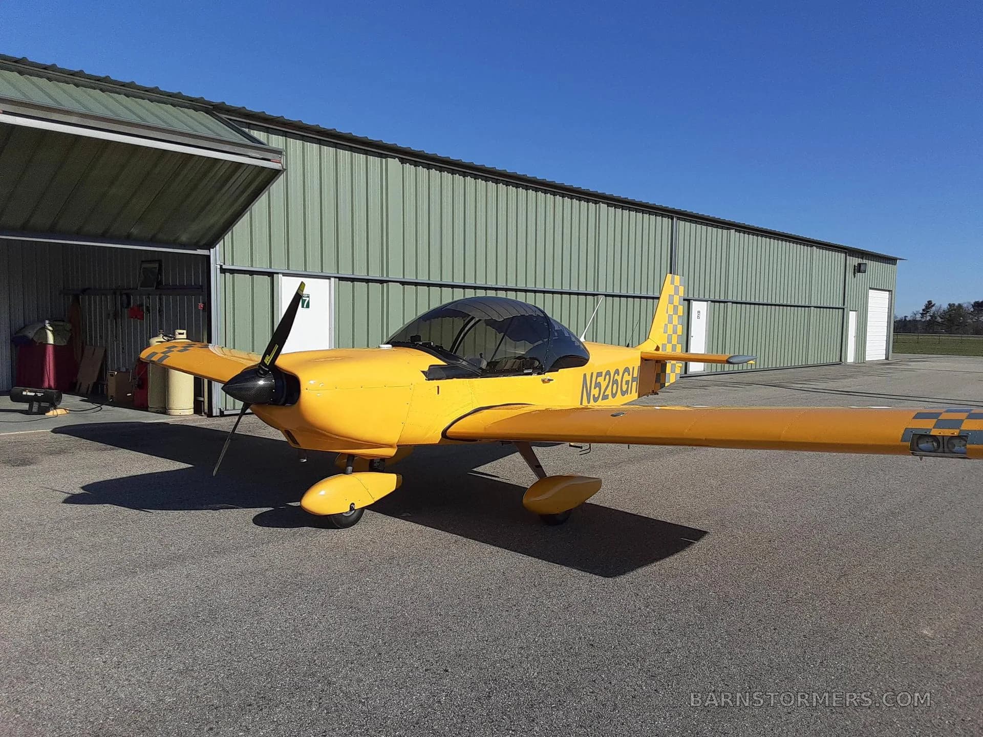 A bright yellow single-engine aircraft with registration N526GH parked on an asphalt ramp in front of a green hangar under a clear blue sky.