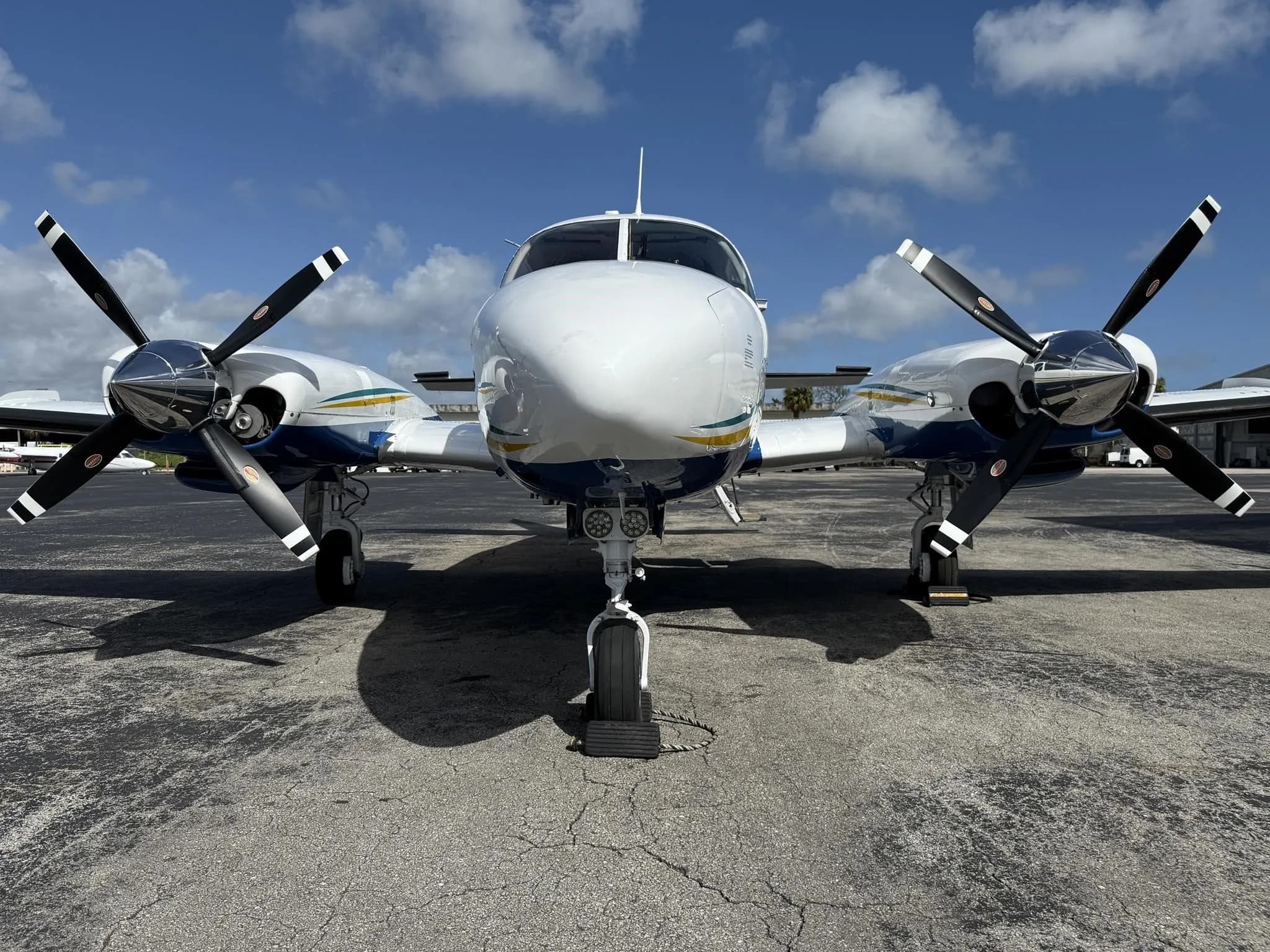 A head-on view of a white and blue twin-engine propeller aircraft with yellow and green stripes, parked on an airport tarmac.