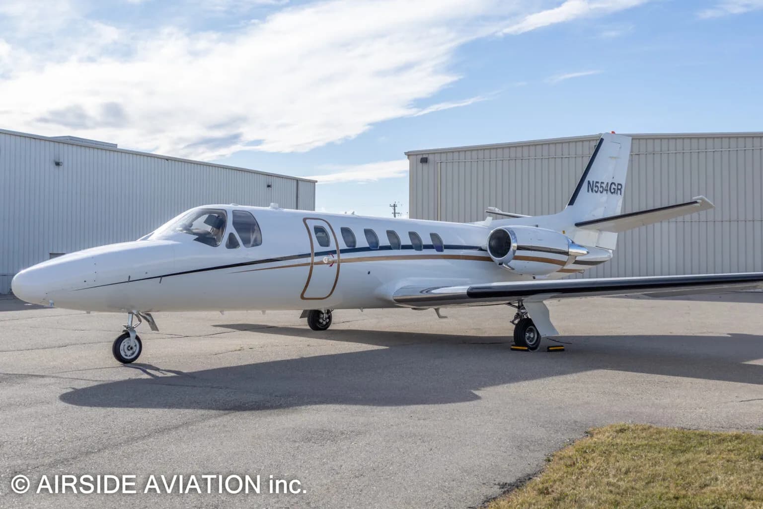 A white Cessna Citation business jet with gold and black stripes, registration N554GR, parked on an airfield.