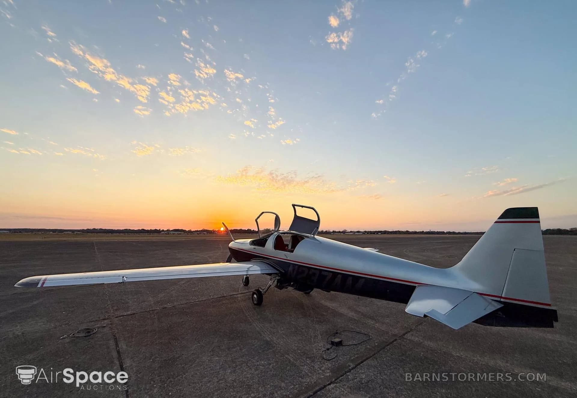 A wide view of a silver and black single-engine aircraft with its canopy doors open, parked on an airfield at sunset.