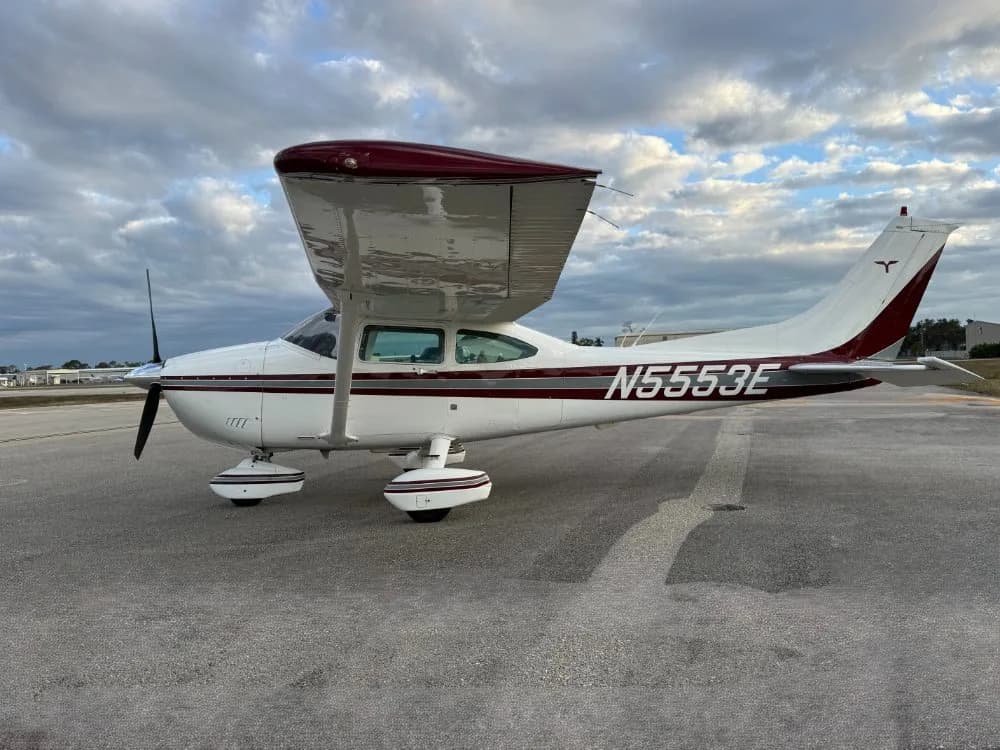 A white and maroon single-engine high-wing aircraft with registration N5553E parked on an airfield under a cloudy sky.