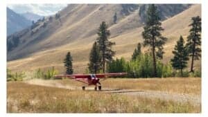 A red Cubcrafters XCub aircraft on a dirt airstrip with mountains in the background.