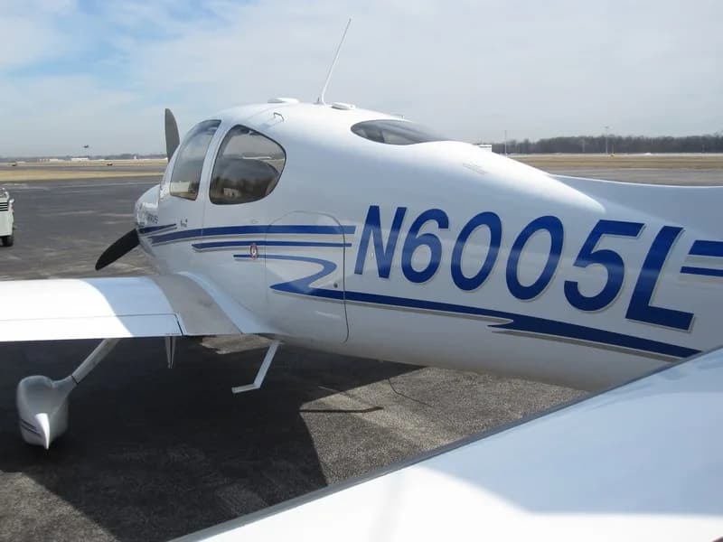 A white Cirrus SR22 aircraft with blue and grey stripes, registration N6005L, parked on an airfield.