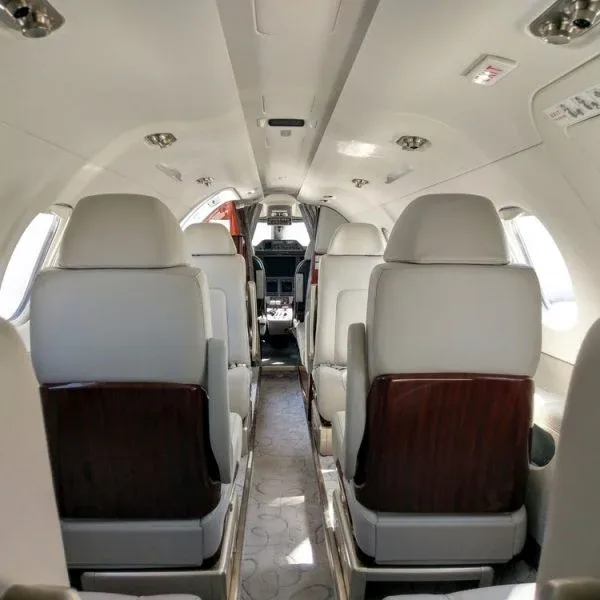 A view down the aisle of a private jet cabin with light grey seats and dark wood accents, looking towards the cockpit.