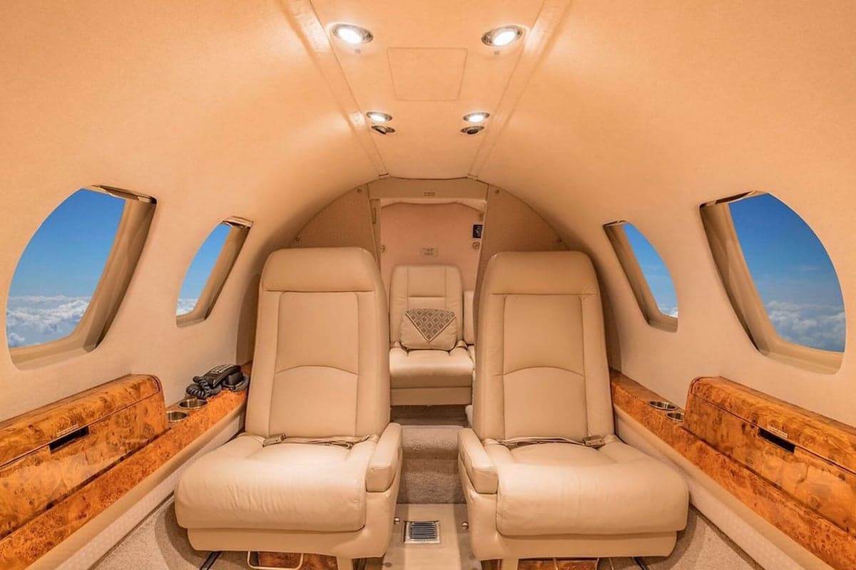 The luxurious cabin interior of a private jet with beige leather seats and wood accents, showing clouds through the windows.