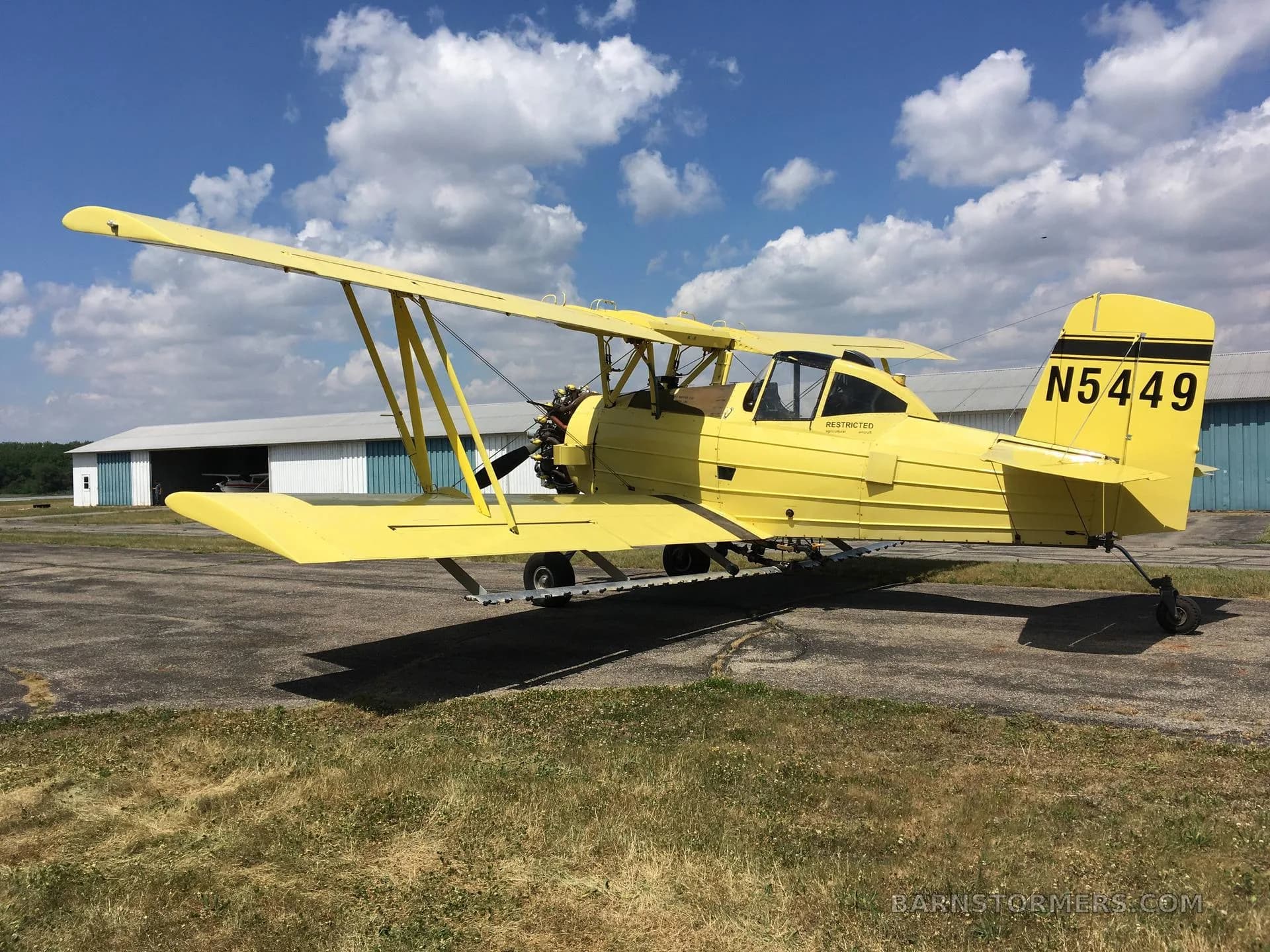 A yellow biplane crop duster, tail number N5449, viewed from the rear-left, parked on an airfield with a white hangar.