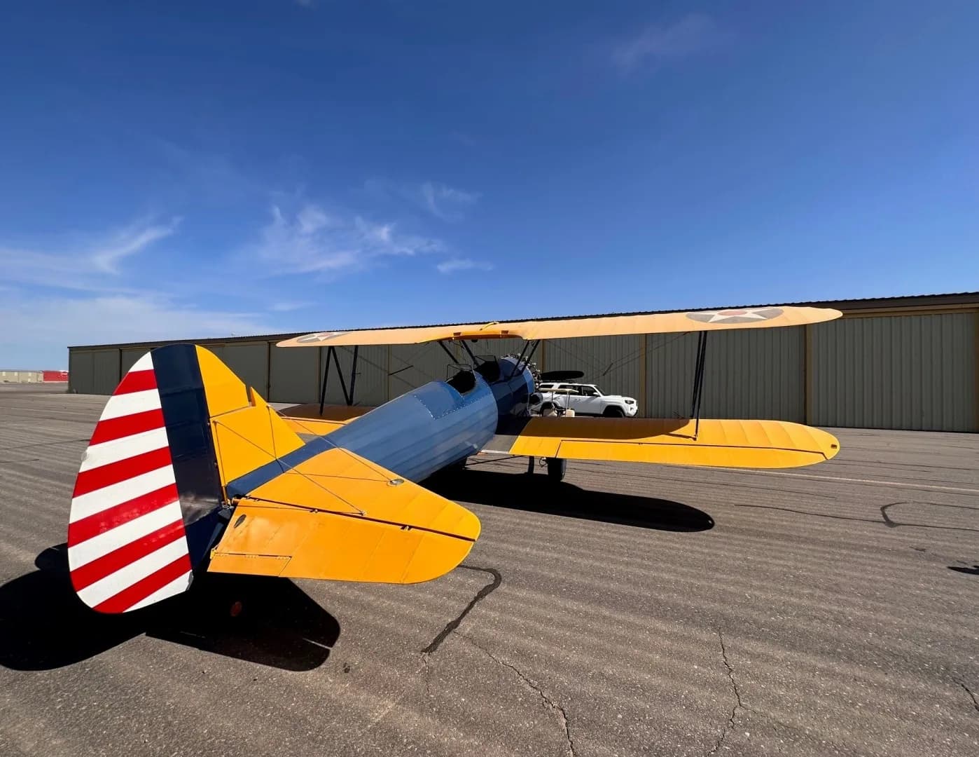 A three-quarter rear view of a blue and yellow biplane with a striped tail, parked on an asphalt tarmac.