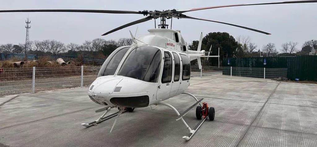 A white Bell 407GXP helicopter with landing skids and ground handling wheels, viewed from the front on a concrete pad.