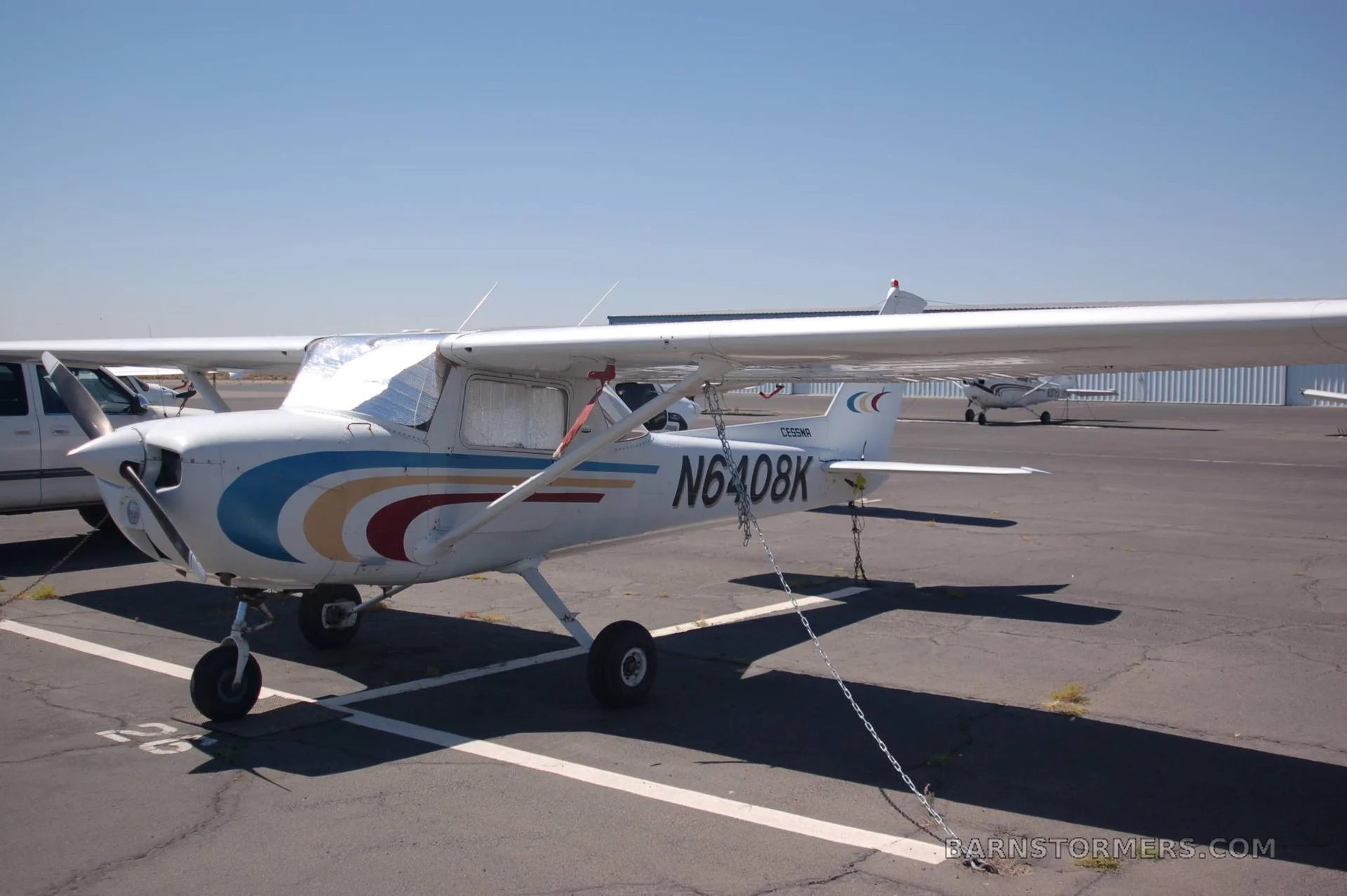 A white Cessna N6408K with blue, gold, and red stripes is parked on an airfield with other aircraft in the background.