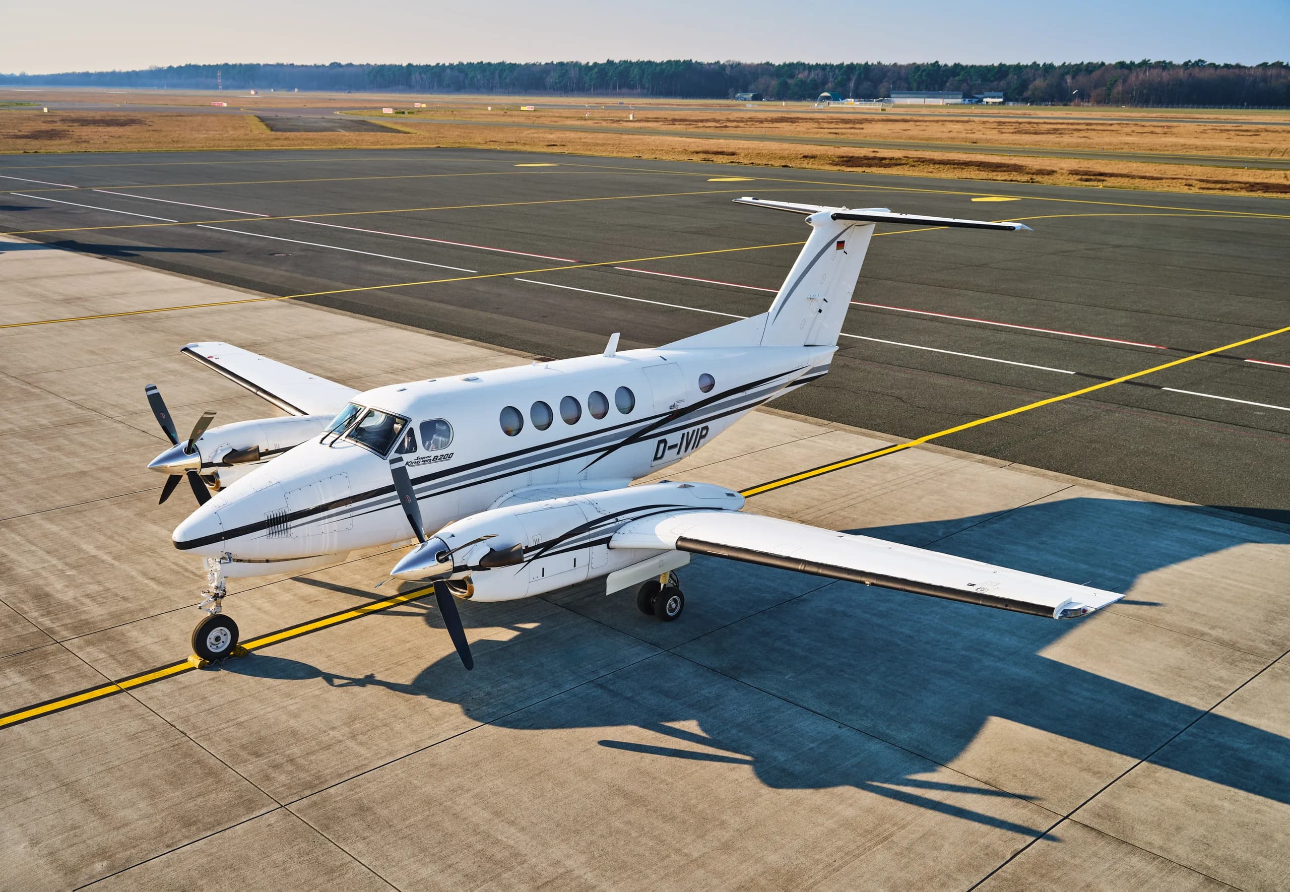 An aerial view of a white and black twin-turboprop aircraft, likely a King Air, parked on an airfield with long shadows.