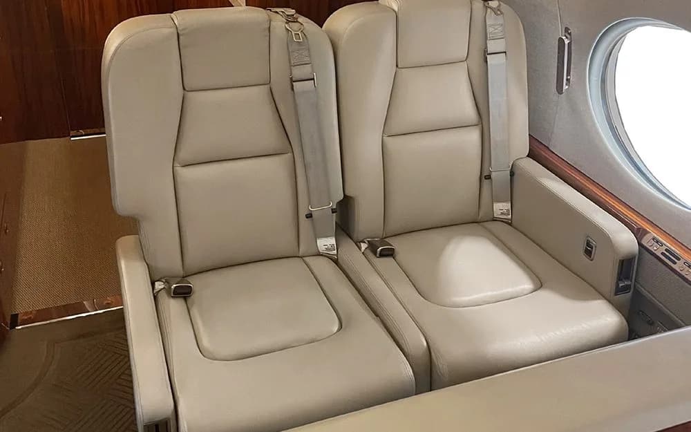 Two light beige leather cabin seats with seatbelts, positioned next to a large window in a private jet interior.