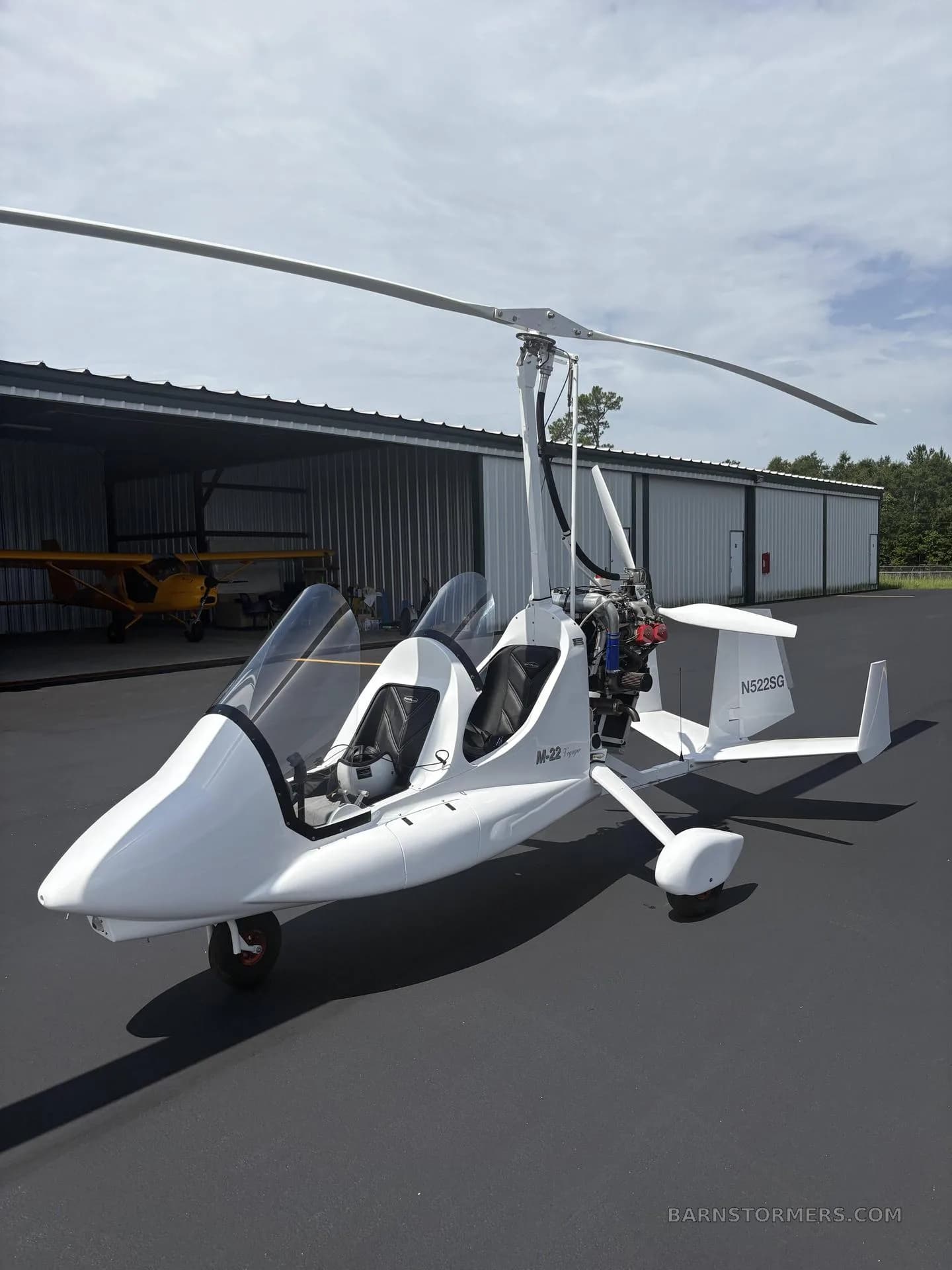 A white M-22 gyrocopter with an open cockpit is parked on an asphalt tarmac with a hangar in the background.