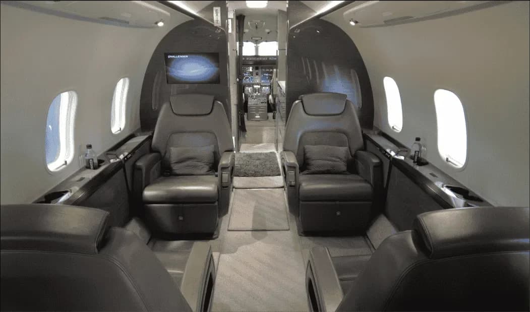 The luxurious interior of a business jet cabin with four grey leather seats facing each other, and the cockpit visible in the background.