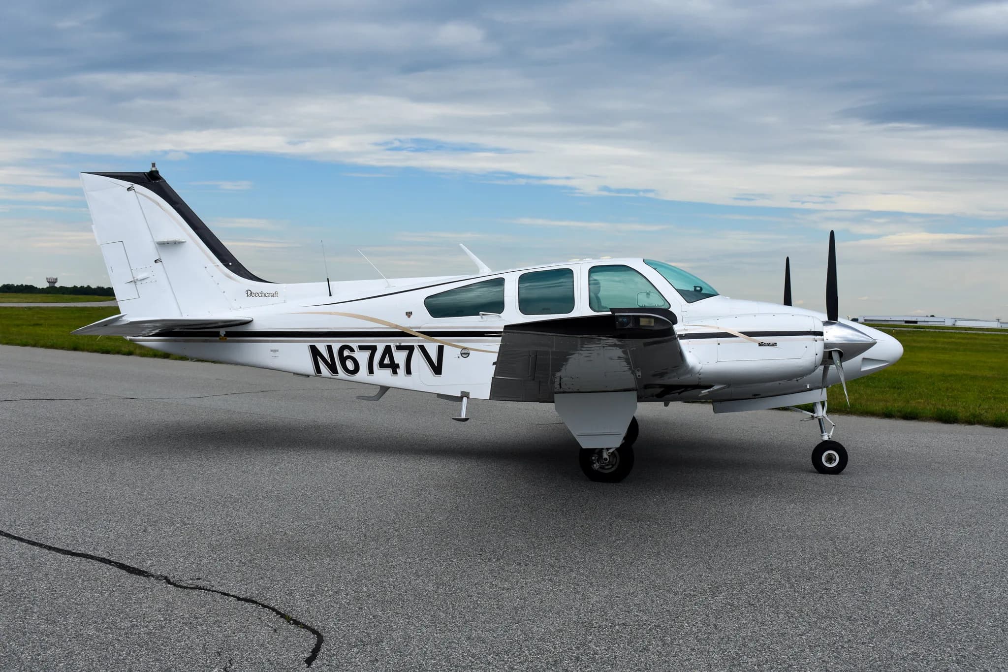 A white and black Beechcraft Baron aircraft, N6747V, is parked on an asphalt runway under a cloudy sky.
