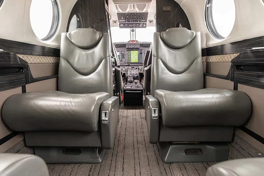 A view from the aircraft cabin looking forward into the cockpit, showing two grey leather passenger seats in the foreground.