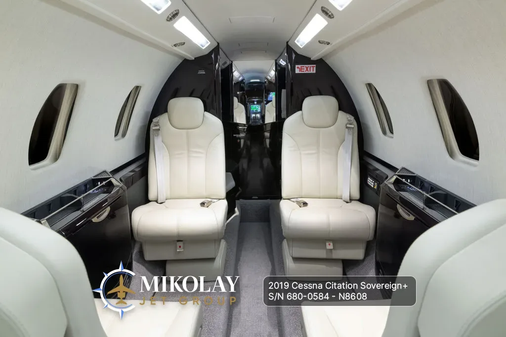 View of the aircraft cabin interior with two rows of light-colored leather seats and dark wood accents, looking towards the front.