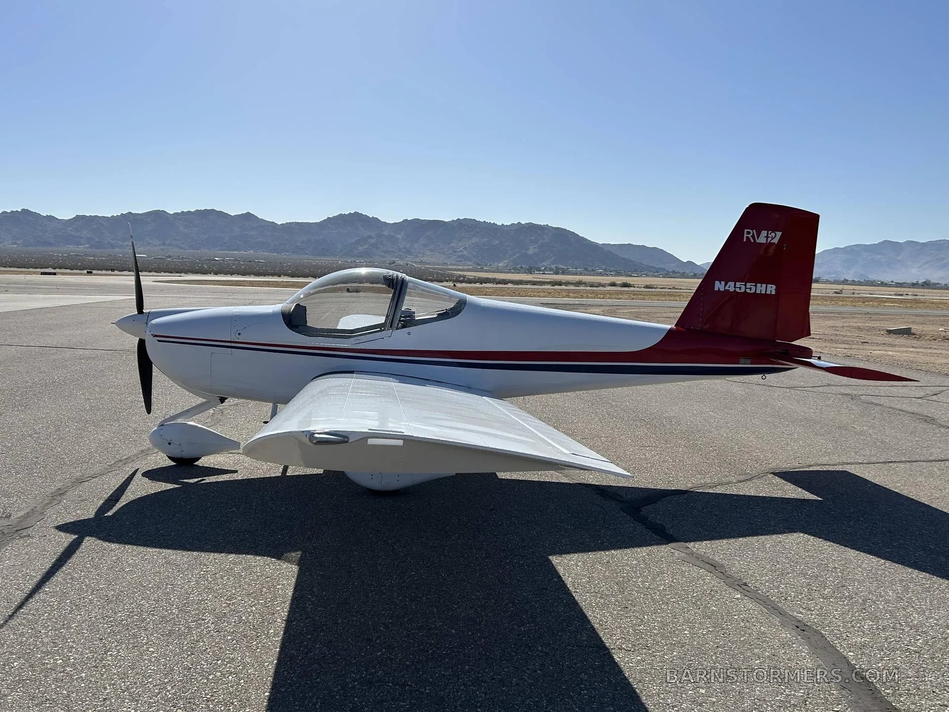A white and red Van's RV-7 aircraft, registration N455HR, is parked on an asphalt tarmac with mountains in the background, viewed from the right side.