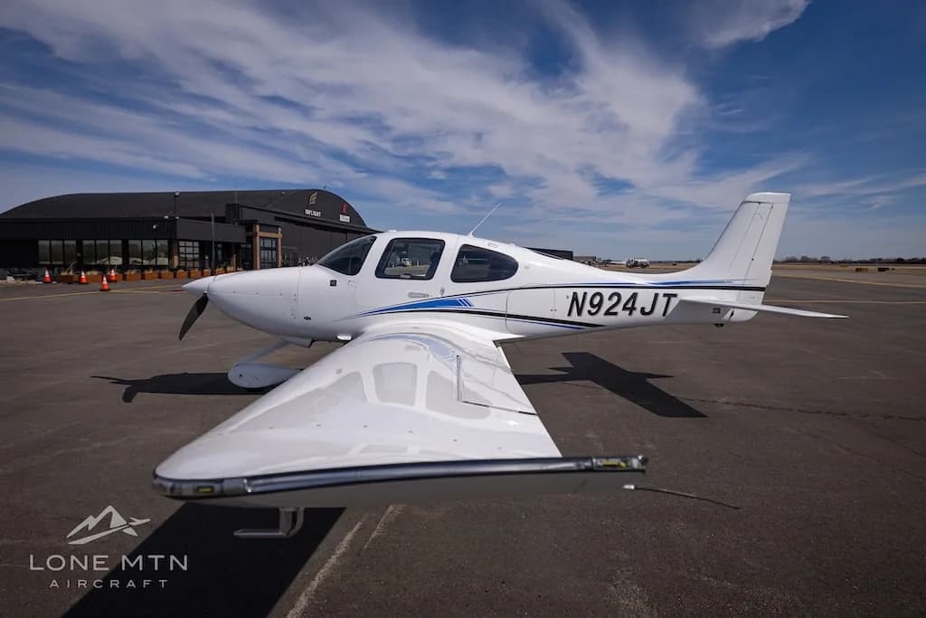 A white single-engine aircraft with blue and black stripes, tail number N924JT, parked on an airport tarmac with a building in the background.