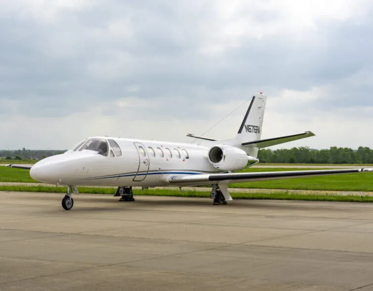 A white Cessna Citation business jet with a blue stripe is parked on an airfield under a cloudy sky.