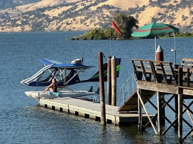 A blue and green seaplane is tied to a wooden dock on a lake with rolling hills in the background.