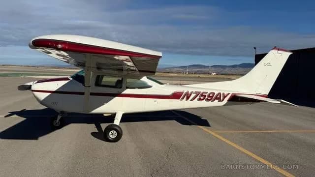 A white Cessna 172 with red and maroon stripes, registration N759AY, parked on an asphalt ramp with mountains in the background.