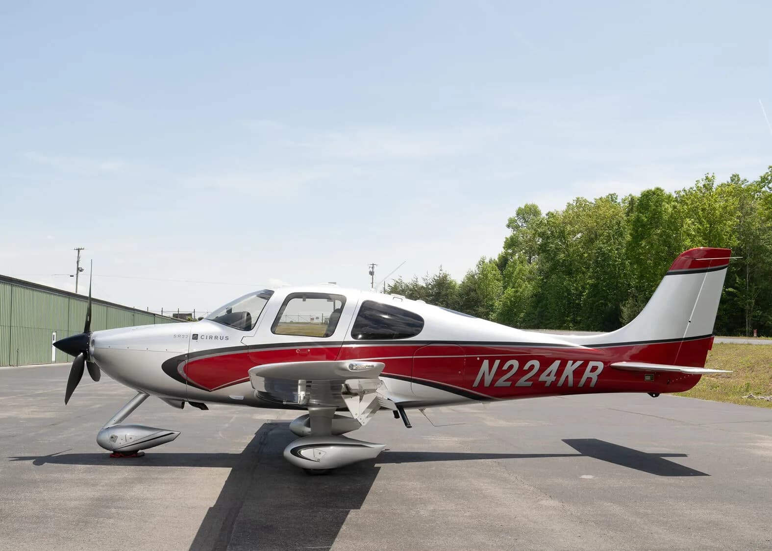 A silver and red Cirrus SR22 aircraft, N224KR, parked on an asphalt ramp with a green hangar and trees in the background.