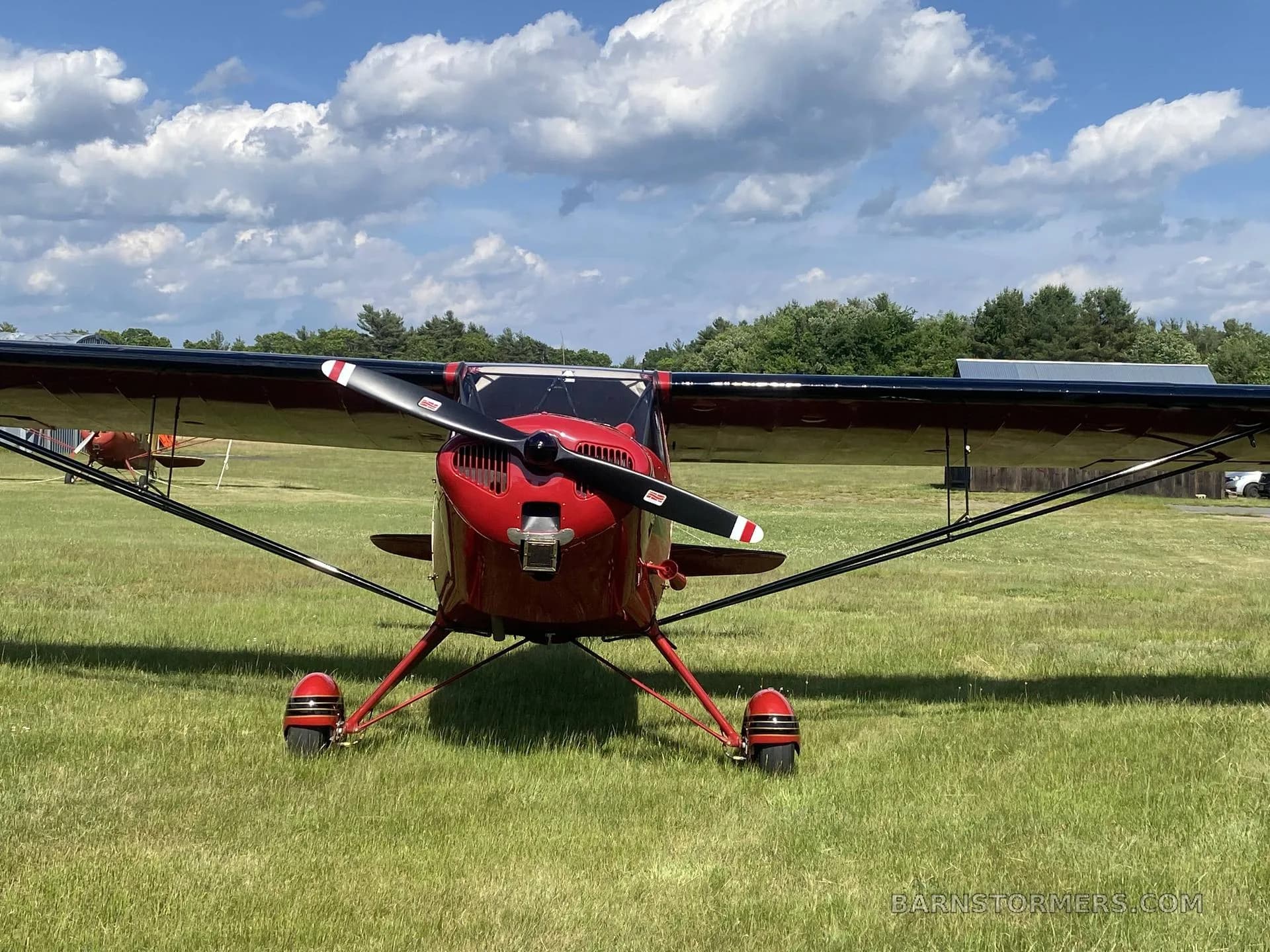 A red single-engine high-wing aircraft with a black propeller, viewed from the front on a grassy airfield.